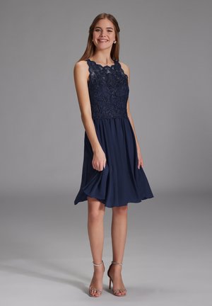 Robe bleu marine avec un corsage en dentelle et broderies florales, jupe évasée et design sans manches. Des sandales à talons argentées complètent le look.