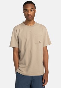 Helles beige T-Shirt mit kurzen Ärmeln aus Baumwolle, ausgestattet mit einer Brusttasche und einem kleinen geometrischen Logo. Glatte Textur und lässige Passform.