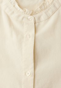 Camicia button-up color crema con colletto arricciato, caratterizzata da strisce verticali e piccoli bottoni bianchi rotondi lungo la parte anteriore.