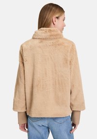 Giacca in pelliccia sintetica beige con colletto alto, spalle scese e polsini a coste, caratterizzata da una superficie morbida e testurizzata e da una vestibilità comoda.
