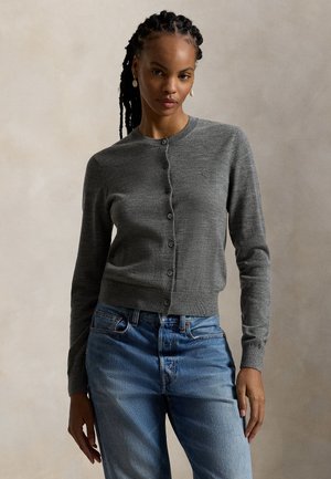 Femme aux cheveux tressés portant un cardigan gris boutonné et un jean bleu, debout devant un fond neutre uni.
