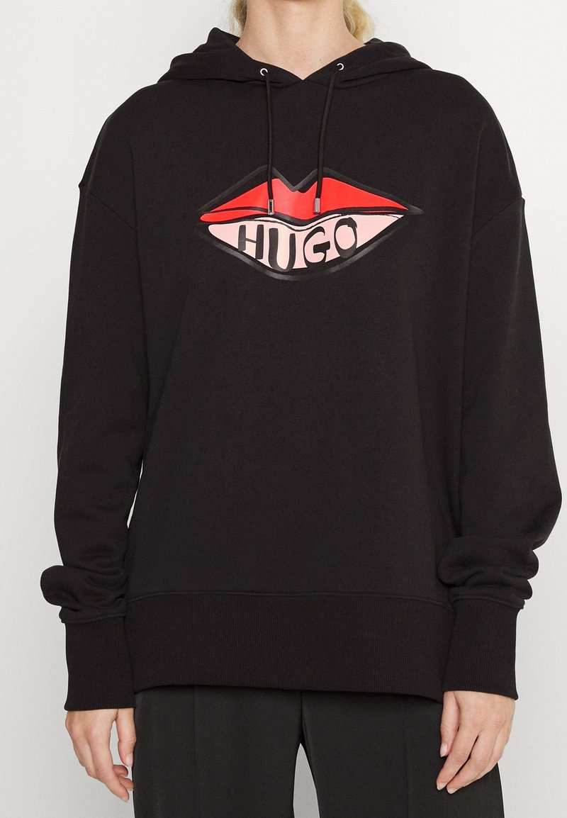 Sweat à capuche noir avec un motif de lèvres rouges et roses affichant « HUGO », porté par une personne avec les mains détendues le long du corps.