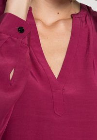 Top fuchsia avec un décolleté en V profond, présentant un léger éclat et un détail de bouton noir sur le poignet. Texture de tissu lisse et léger.