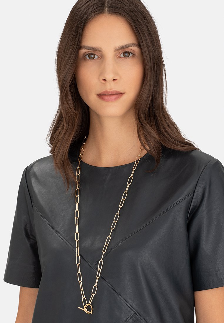 Sort læder T-shirt med korte ærmer med et geometrisk design. Et langt guldkæde med ovale led og en cirkulær lukning tilføjer detaljer.