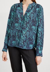 Blouse à manches longues de couleur bleu marine avec des motifs tourbillonnants en teal et noir, présentant un décolleté en V profond et une coupe décontractée. Tissu lisse et léger.