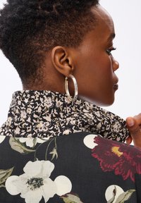 Profil d'une personne avec des cheveux courts et bouclés, portant de grandes boucles d'oreilles et une chemise noire à motif floral avec un col relevé.