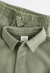 Hellgrünes Strickpolo mit Kragen und Knopfverschluss, kombiniert mit Hosen mit elastischem Bund aus demselben Stoff und in der gleichen Farbe.