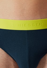 Tummansininen alushousu, jossa on teksturoitu keltainen vyötärönauha. Vyötärönauha on varustettu kohokuvioidulla brändin nimellä "Schiesser". Materiaali näyttää pehmeältä ja hyvin istuvalta.