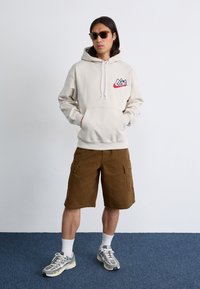 En ung mand står selvsikkert i en beige hoodie og brune cargo-shorts, iført solbriller og stilfulde sneakers imod en ensfarvet baggrund.
