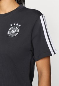 Sötétszürke pólót viselő személy, amelyen a Német Labdarúgó Szövetség logója található négy csillaggal a felette, és fehér csíkok láthatók az ujjon.