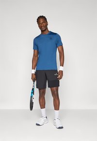 Sininen urheilupaita, mustat shortsit, valkoiset rannekkeet, käsissään tennismaila, valkoiset sukat ja lenkkarit. Sujuva, rento muotoilu.