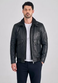 Man draagt een zwarte leren jas over een wit T-shirt en donkere broek, staat met één hand in zijn zak tegen een effen achtergrond.