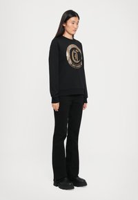 Schwarzer Hoodie mit einem runden Logo aus goldenen Pailletten, kombiniert mit schwarzen Schlaghosen und klobigen schwarzen Stiefeln. Glatte Textur, lässiges Design.