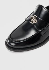 Mocasini din piele lacuită neagră, cu o cataramă argintie decorativă cu monogramă, încrustată cu rhinestones pe partea superioară lustruită.