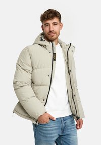 Jasno beżowa kurtka puchowa z kapturem, z pikowaną fakturą, zamkiem błyskawicznym z przodu i napami, noszona na białym swetrze i dżinsach.