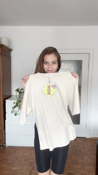 Kermanvärinen lyhythihainen t-paita pyöreällä kaula-aukolla, jossa on printattu logo ja tarrat. Sileä tekstuuri, rento malli.