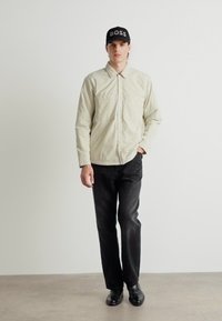 Light beige button-up shirt with two front pockets, paired with black straight-leg trousers and black shoes, topped with a black cap.