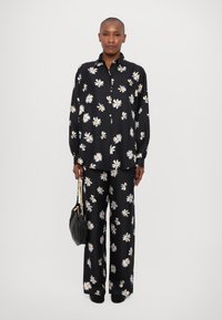 Cămașă cu un model floral pe fond negru și pantaloni largi, cu margarete albe. Materialul are o textură fină. O geantă neagră este ținută într-o mână.