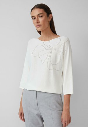 Maglione bianco con trama, a tre quarti di manica, caratterizzato da un design floreale outline in cuciture argentate. Abbinato a pantaloni grigi.