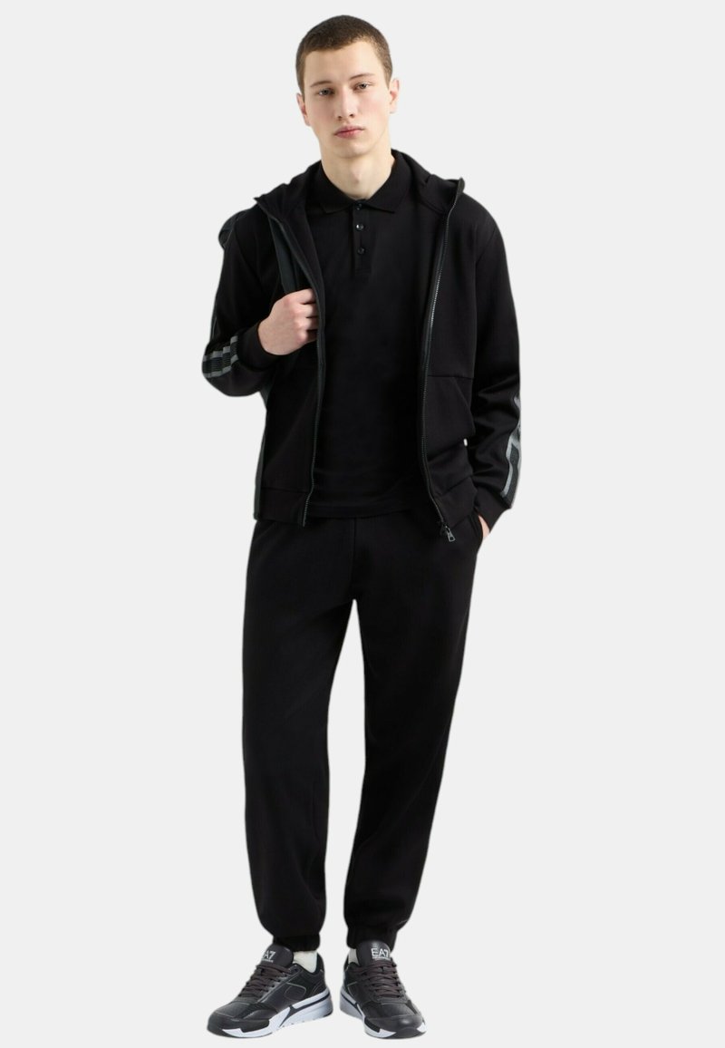 Felpa nera con zip e motivo a scacchi sulle maniche, abbinata a una polo nera e pantaloni della tuta coordinati. Indossata con scarpe da ginnastica grigie.