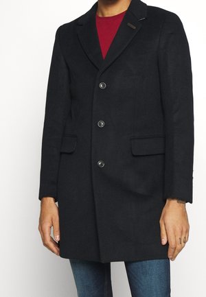 Homme portant un manteau noir boutonné sur une chemise rouge et un jean bleu, debout les mains détendues le long du corps.