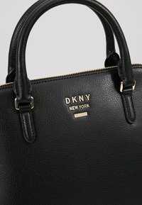Černá kožená kabelka s texturovaným povrchem, zlatým kováním, dvojitými uchy a vyraženým logem "DKNY NEW YORK." Zipový uzávěr na vrchu.