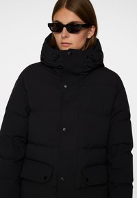 Svart puffjacka med hög krage, framknappar och sidofickor. Slät tyg med en quiltad textur.