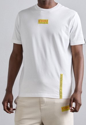 T-shirt bianco in cotone con logo ricamato giallo "ICEBERG" e due etichette con testo giallo aggiuntive sul davanti e sul lato.