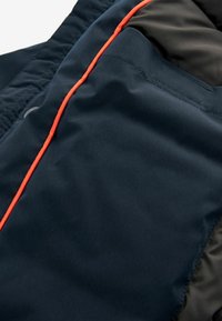 Giacca isolante blu scuro con vestibilità slim, texture liscia, piping arancione e tasca nascosta sul lato sinistro.