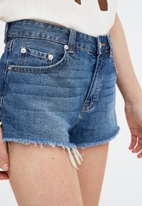 Denim-shortsit keskisinisellä pesulla, joissa on rispaantunut helma, etutaskut ja hopeiset nappikoristeet. Vakioistuvuus ja korkea vyötärö.