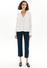 Blusa blanca de manga larga con escote en V y detalle plisado, combinada con jeans cortos de color azul oscuro y mocasines negros. Textura suave en toda la prenda.