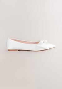 Zapato plano blanco en punta con un gran lazo decorativo en la puntera y forro interior rosa claro sobre un fondo neutro.