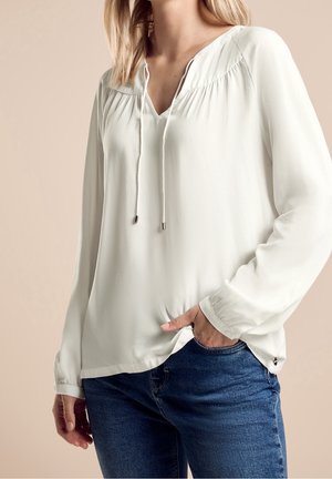 Vrouw draagt een witte blouse met lange mouwen, V-hals en koordjes aan de voorkant, gecombineerd met een blauwe spijkerbroek, hand in de zak, beige achtergrond.