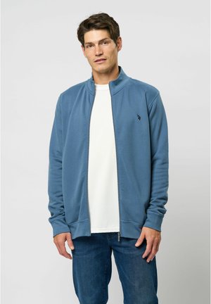 Blå zip-up jakke lavet af blødt stof, med høj krave, ribbede manchetter og et lille logo på brystet, parret med en hvid skjorte.