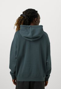 Felpa con cappuccio oversize in teal scuro, realizzata in tessuto morbido. Presenta un cappuccio ampio e polsini a costine, con una vestibilità comoda e una texture tattile.