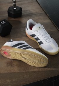 White training shoes with black stripes, textured upper, gum sole, and red accent. Features a padded collar and lacing system.