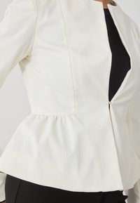 Fehér peplum zakó sima textúrával, rövid álló gallérral és középső cipzárral. Fekete anyag látható alatta.
