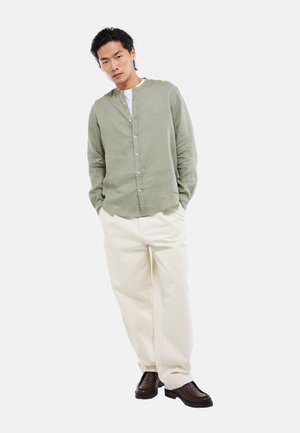 Homme debout, les mains dans les poches, portant une chemise boutonnée vert clair, un pantalon blanc et des chaussures marron foncé sur fond blanc.