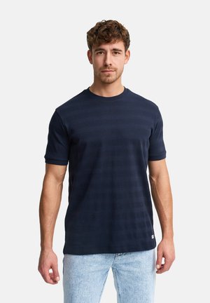 Mężczyzna o krótkich kręconych włosach, ubrany w granatową, paskowaną koszulę z krótkim rękawem oraz jasnoniebieskie dżinsy, stojący na jednolitym tle.