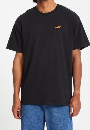 Mężczyzna w gładkiej, czarnej koszulce z krótkim rękawem z małym pomarańczowym logo "Volcom" na piersi oraz w niebieskich dżinsach.