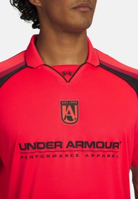 Muž v jasne červenom polo tričku Under Armour s čiernymi akcentmi, logom na hrudi a textom "Performance Apparel" pod ním.