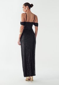 Črna večerna obleka z odprtimi rokavi brez naramnic, okrašena s svetlečimi paličicami, oprijetim krojem in dolžino do tal. Ima nizko hrbtno stran.