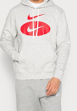 Man draagt een lichtgrijze Nike-hoodie met een groot rood dubbele swoosh-logo en bijpassende grijze trainingsbroek met wit Nike-logo.