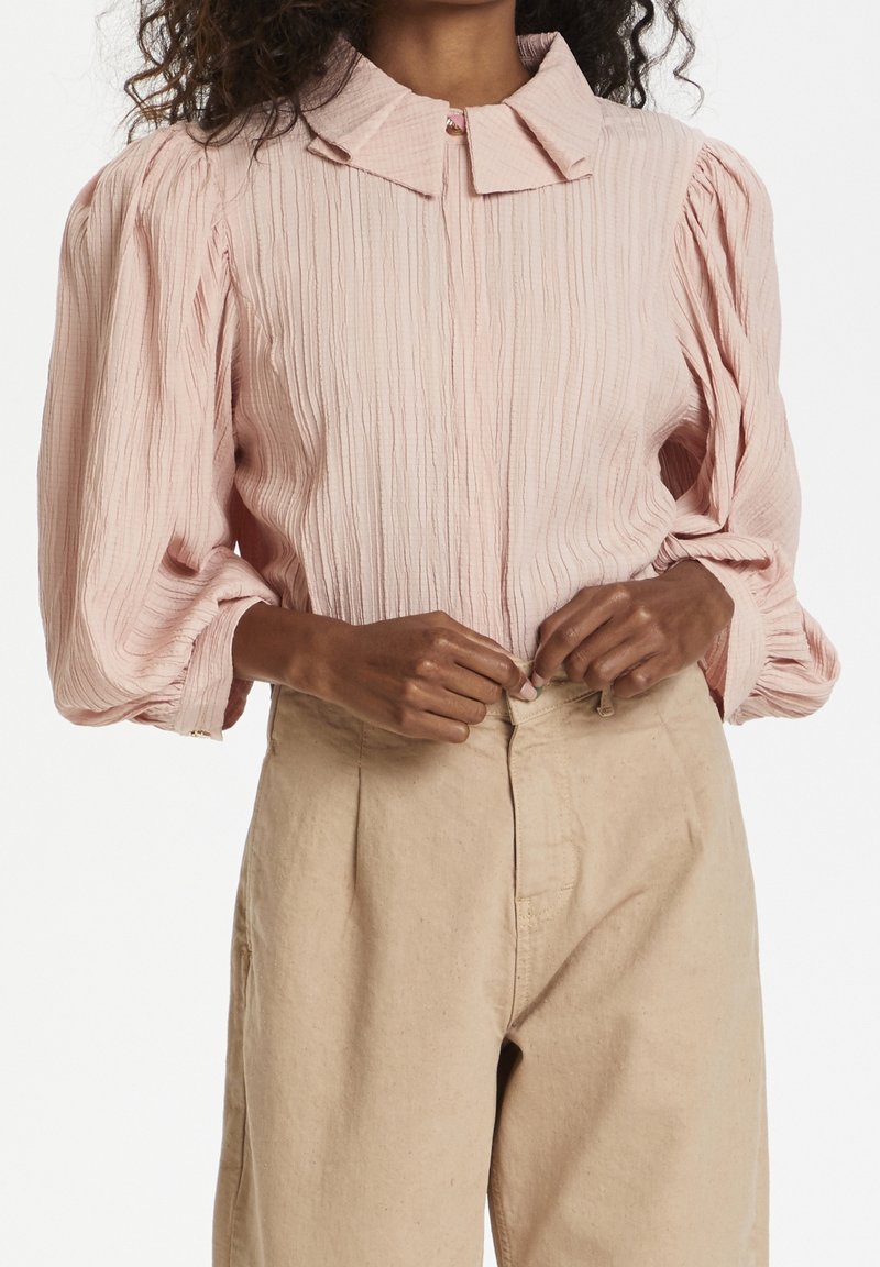 Femme portant un chemisier texturé rose pâle avec des manches bouffantes et un pantalon beige taille haute, en train de boutonner un bouton à la taille.