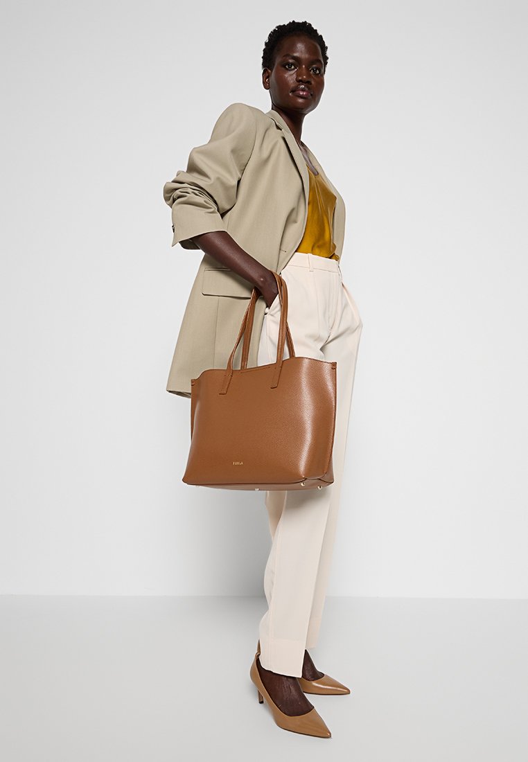 Brązowa skórzana torba tote z podwójnymi uchwytami, w strukturalnym kształcie, z subtelnym wytłoczonym logo. Model wystylizowany w beżową marynarkę i kremowe spodnie.