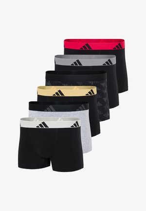 Gut gebauter dunkelhäutiger Mann steht barfuß und trägt schwarze Boxer-Shorts mit einem breiten gelben Bund und Adidas-Logos, schaut zur Seite.