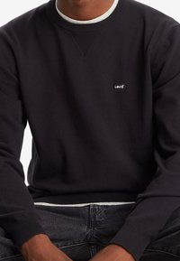 Svart crewneck sweatshirt med ribbad fåll och manschetter, med en liten vit "Levi's"-logotyp på bröstet. Mjuk textur och avslappnad passform.