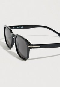 Μαύρα γυαλιά ηλίου με γεωμετρικό πλαίσιο, φακούς με γκρι απόχρωση και μεταλλικές λεπτομέρειες στα πλαϊνά. Το όνομα της μάρκας "PIER ONE" στο μπράτσο.