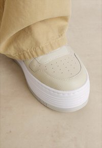 Sneaker piattaforma bianca con tomaia liscia e strutturata, dettagli perforati e una suola in gomma spessa. Abbinata a pantaloni beige chiaro.