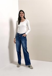 Kvinde står iført hvid skjorte med lange ærmer, blå vidde jeans og sneakers, med den ene hånd i lommen mod en ensfarvet baggrund.